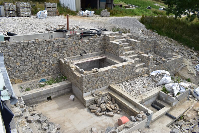 Stone-walled construction site showing partially built structure with dry stone walls, steps, and foundation work in progress