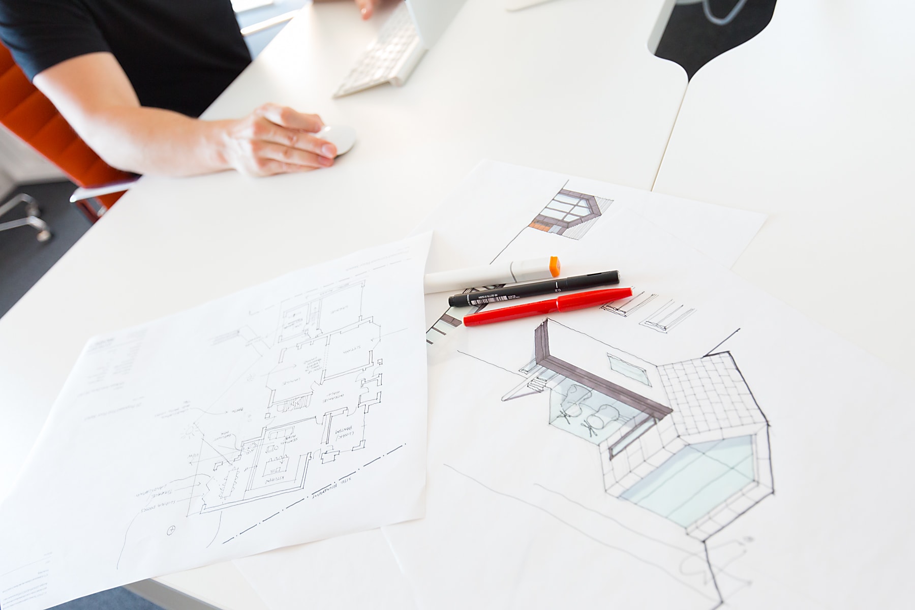 Designer's hand using a computer mouse beside architectural floor plans and a hand-drawn pool elevation sketch