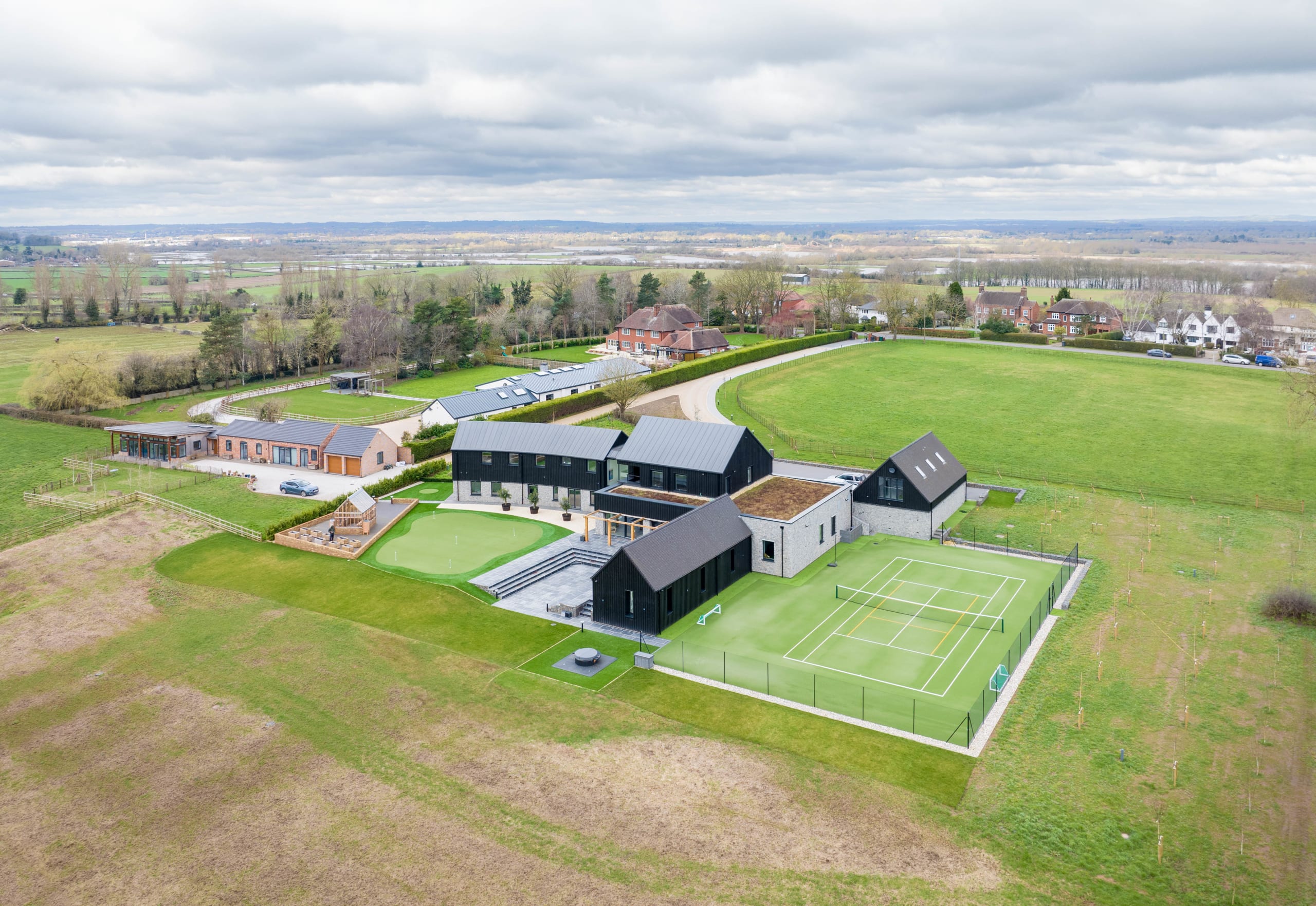 Aerial view of a black-clad barn-style rural residence with tennis court, putting green, and stables amid open farmland