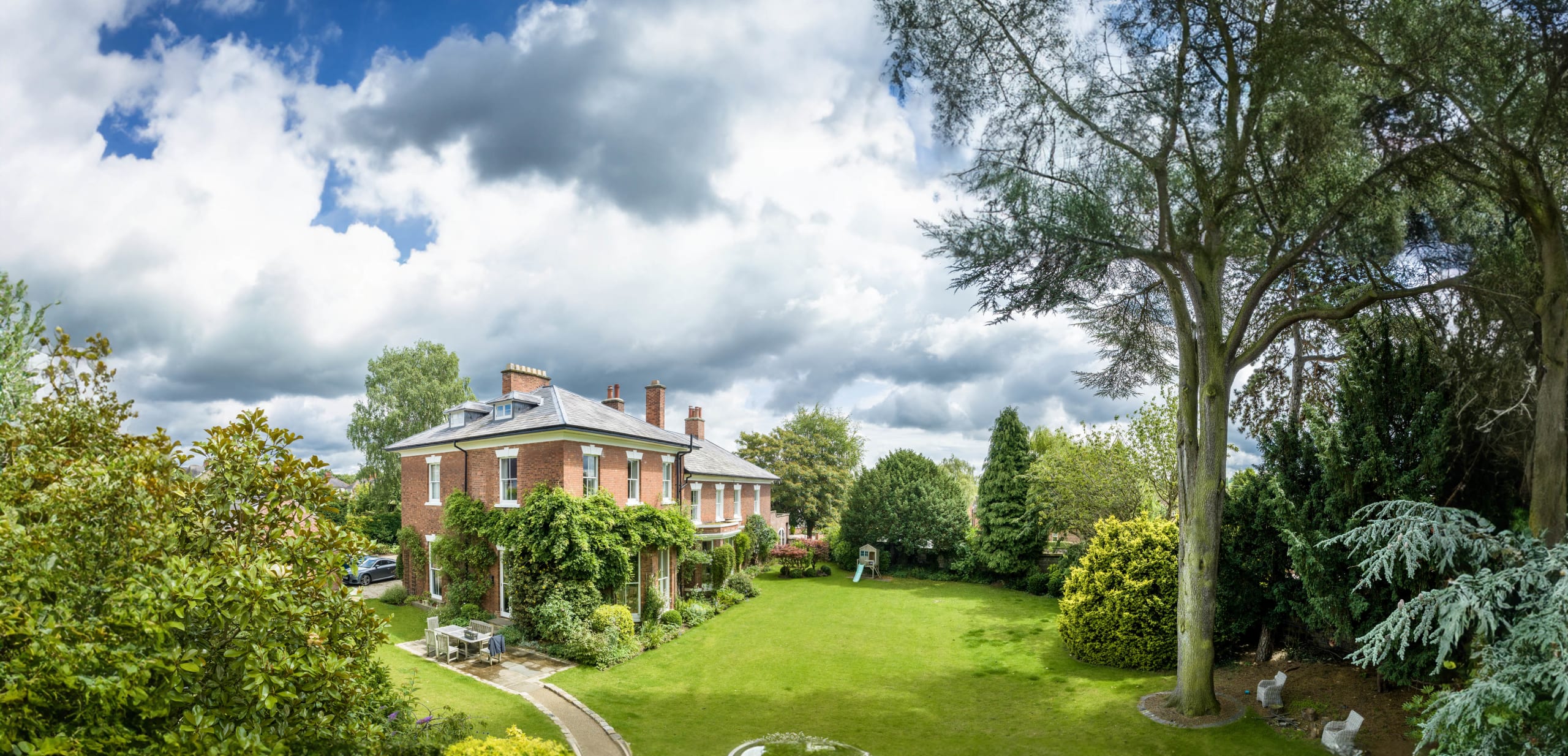 Georgian red brick mansion with slate roof surrounded by manicured gardens and mature trees under dramatic cloudy sky
