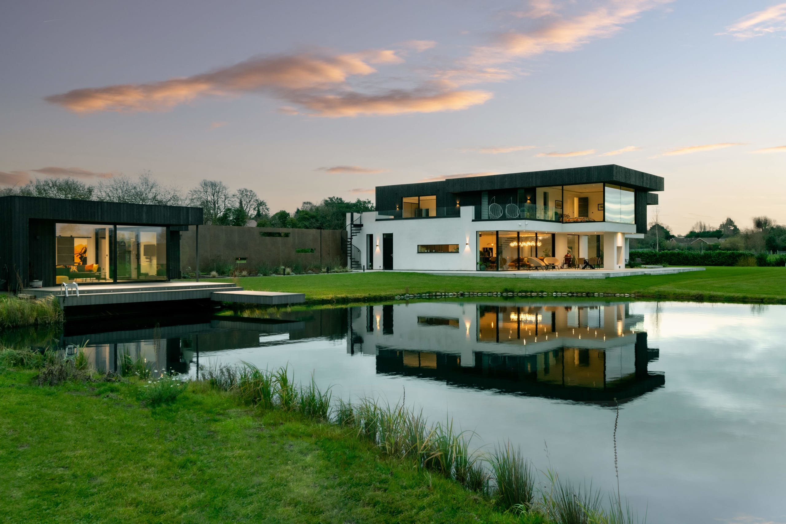 Modern two-story white and black home with glass walls and separate pool house reflected in pond at dusk