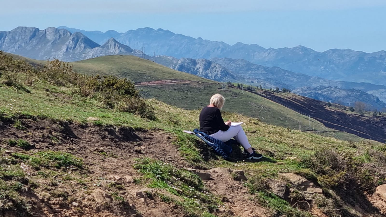 A woman sitting on a hillside sketching, the jagged Picos de Europa behind