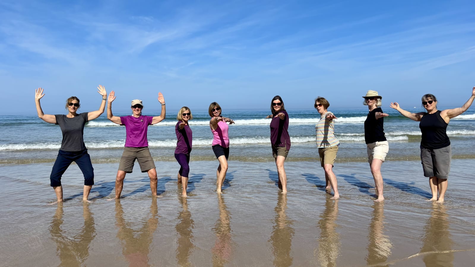 Eight women at the water's edge with their arms raised, reflected in the wet sand