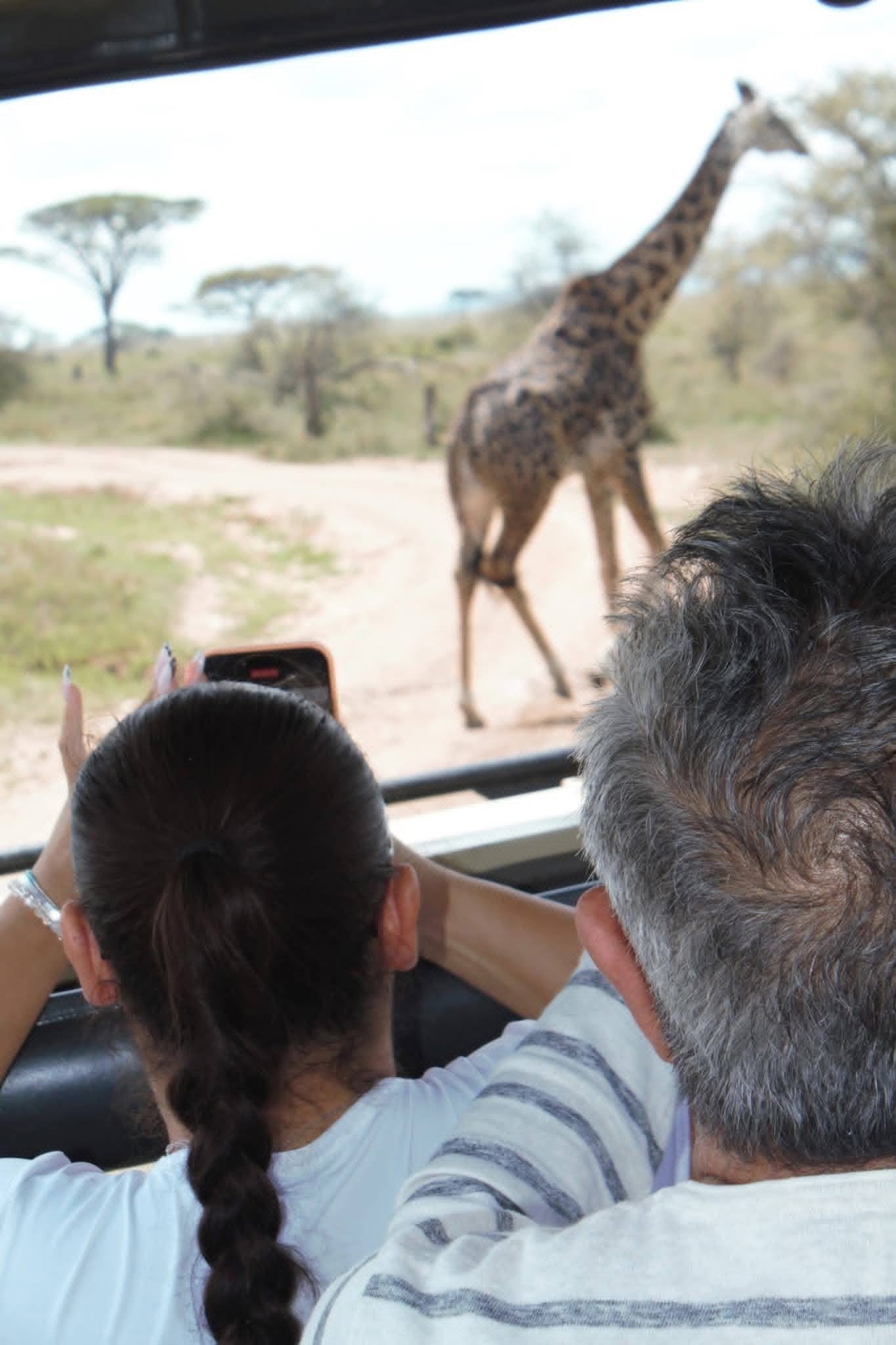 Safari guests photographing a giraffe up close from a private game drive vehicle in Tanzania
