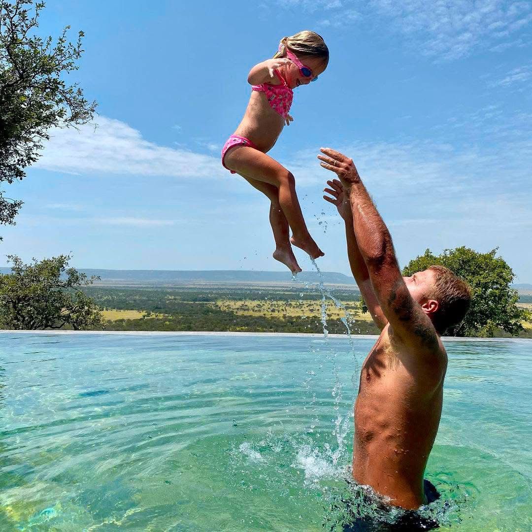 Children swimming in a safari lodge pool in Tanzania, with acacia trees and African bush in the background during midday break