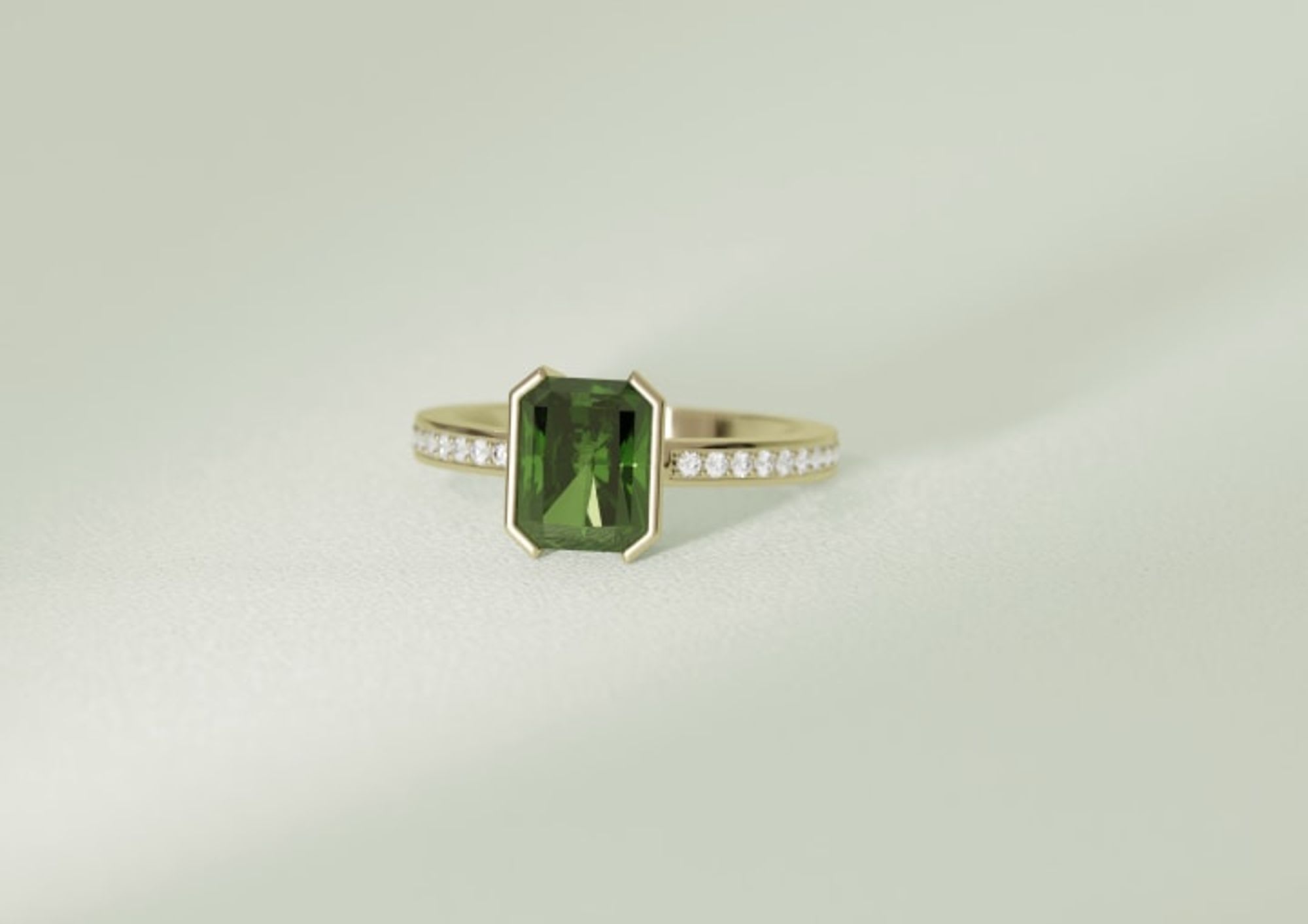 Green solitaire emerald diamond held by two half bezels with a gold band and channel set white diamonds