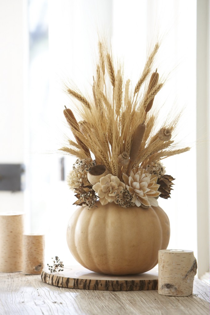 Dried foliage springs forth from a base of off-white flowers and a synthetic pumpkin. Wheat, dried cattails, and dried poppy blooms include height to the center of the display screen. Silk flowers call this center display screen, peaking out of the off-white pumpkin base. This round focal point works well on smaller sized table or sideboards.