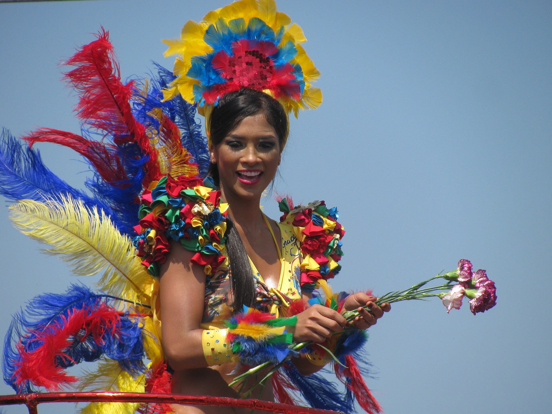 Bailarina con tocado de plumas amarillas, azules y rojas y flores en la mano durante un desfile del Carnaval de Barranquilla