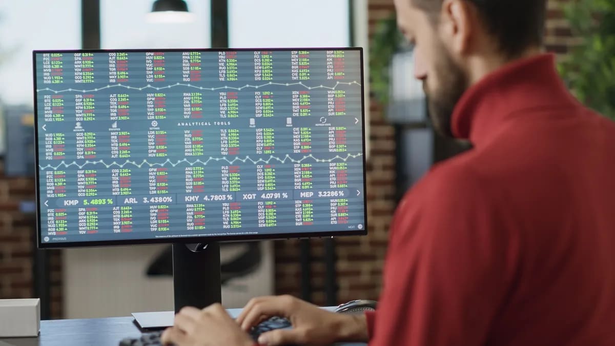 Hombre de suéter rojo que trabaja frente a un monitor con tablas de cotizaciones