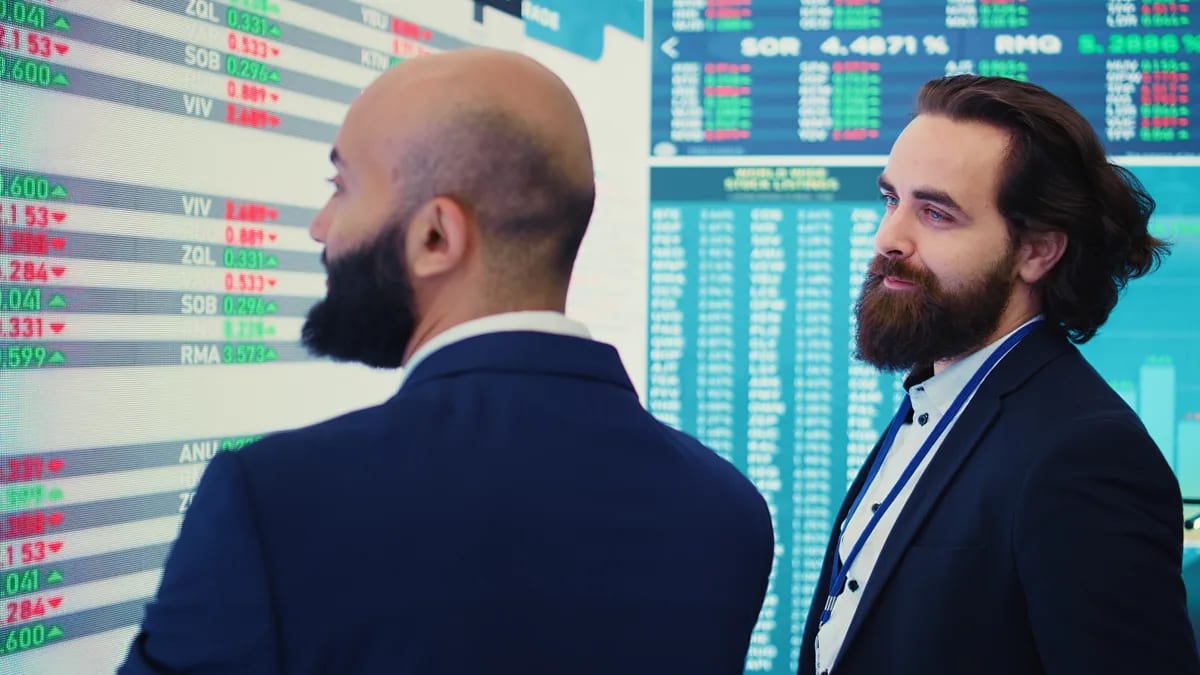 Dos hombres de traje conversan frente a un tablero de cotizaciones