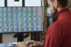 Hombre de suéter rojo que trabaja frente a un monitor con tablas de cotizaciones