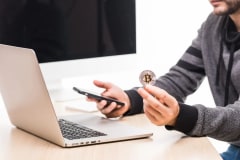 Hombre que sostiene una moneda de Bitcoin y un celular frente a una laptop