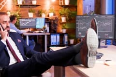 Hombre de traje al teléfono con los pies sobre el escritorio, frente a monitores con gráficas