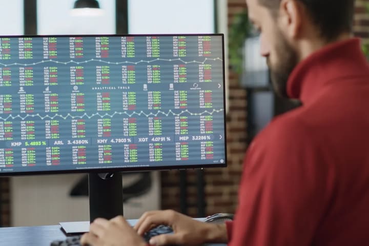 Hombre de suéter rojo que trabaja frente a un monitor con tablas de cotizaciones