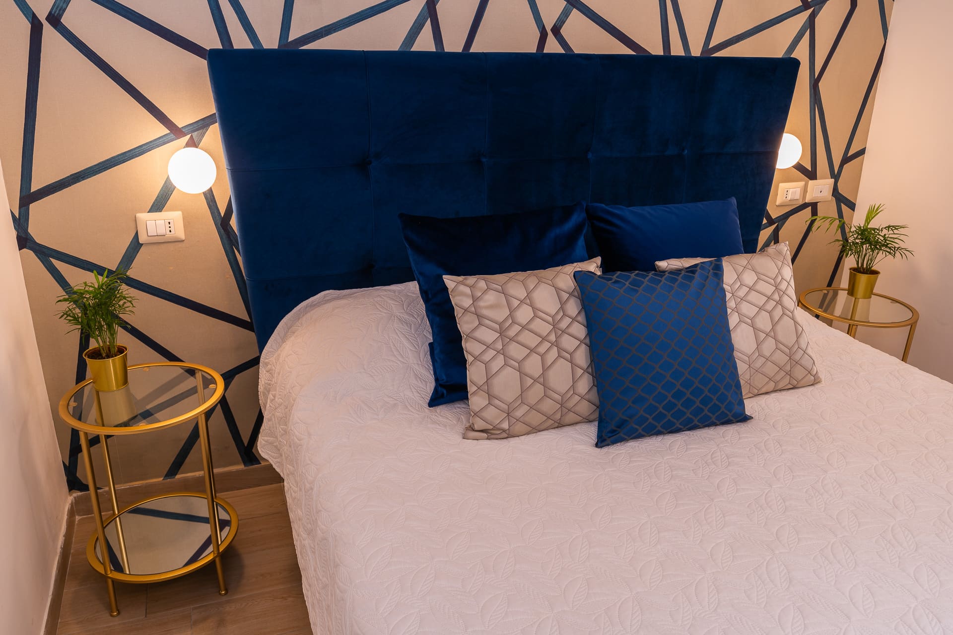 Testiera in velluto blu della camera Megaride con parete geometrica, comodini dorati e cuscini decorativi
