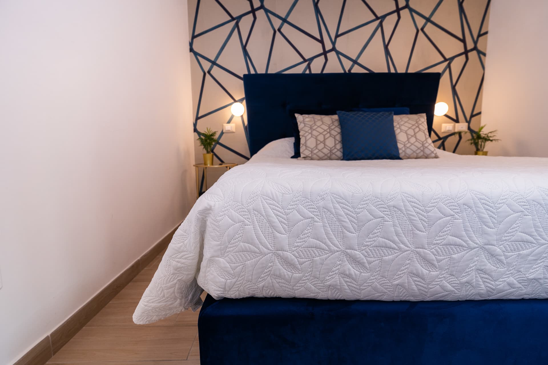 Letto matrimoniale della camera Megaride con copriletto trapuntato bianco e parete geometrica blu e oro