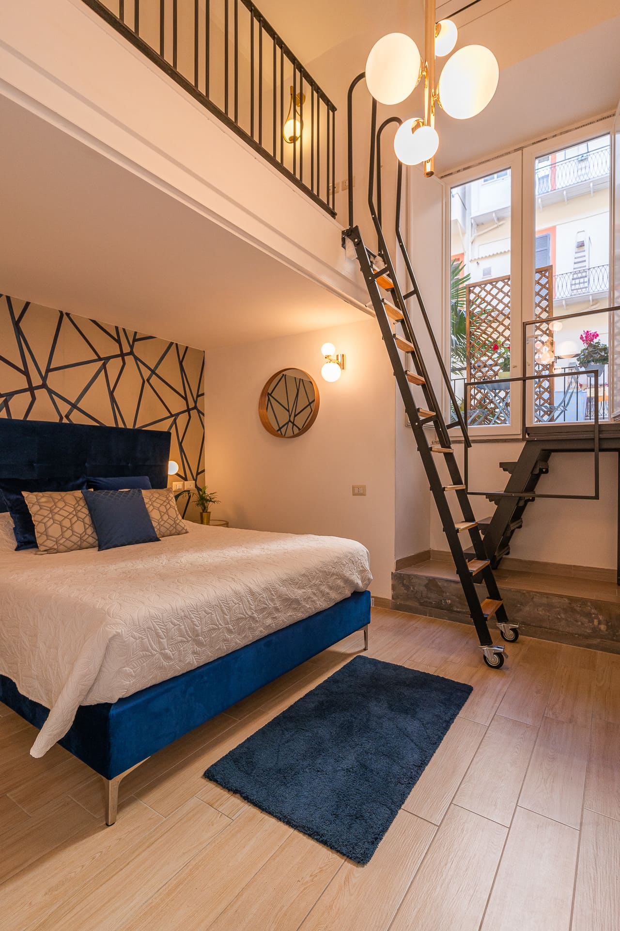 Camera Megaride: letto matrimoniale con testiera in velluto blu e scala in ferro verso il soppalco, Daymoon House Vanvitelli