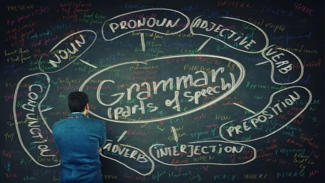 A boy is standing near a green chalkboard with English grammar examples