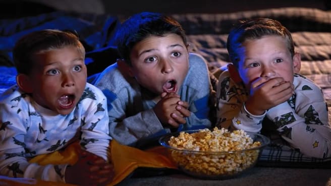 Three funny kids watching a movie in their pajamas and eating popcorns.