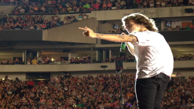 Harry Styles singing on a stage with the crowd in the background.