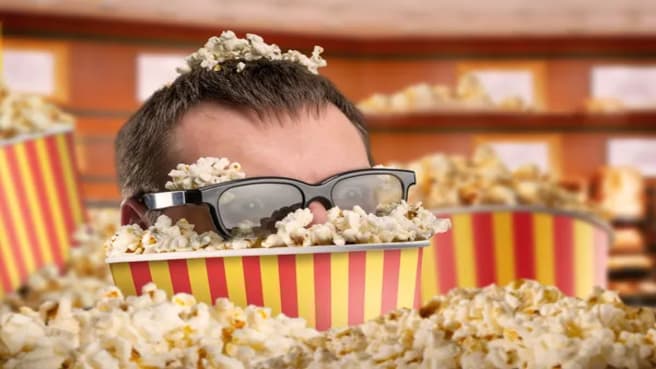 Man in glasses in a bucket of popcorn watching a movie.