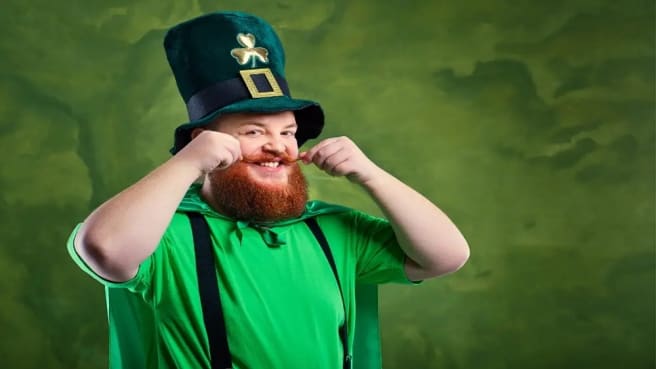 A smiling red-bearded man dressed in green for St. Patrick holding his mustache.