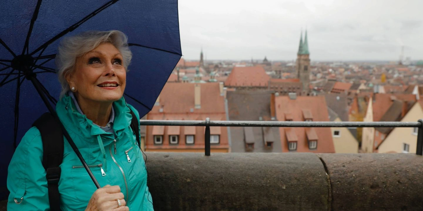 An older woman in a teal jacket holding a blue umbrella and looking out over a European city with red tiled rooftops.