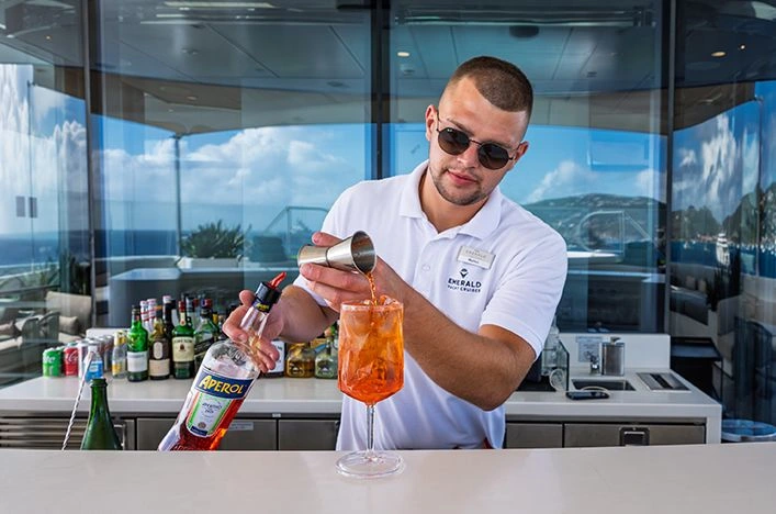 Emerald Azzurra Sky Bar bartender pouring an Aperol Spritz at the outdoor counter, with Caribbean sea views reflected in the glass windows behind.