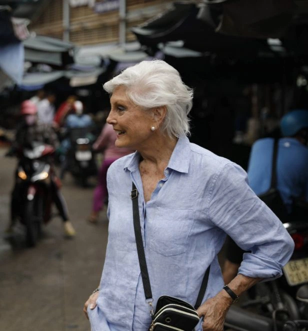 A woman with short white hair wearing a light blue button-down shirt stands in a busy Asian market street.