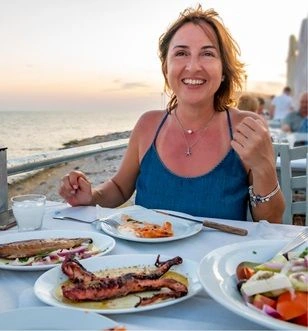 Woman enjoying Greek food and ouzo during sunset with a backdrop of the coastline