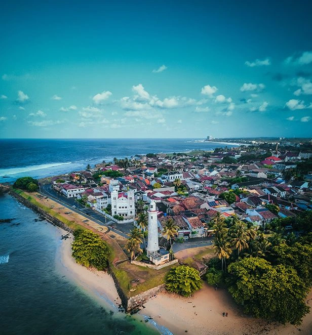 Aerial view of Galle Fort in Sri Lanka, showing a white lighthouse on a grassy promontory, sandy beach and turquoise sea on one side, and a cluster of colorful colonial-style buildings under a bright blue sky on the other.