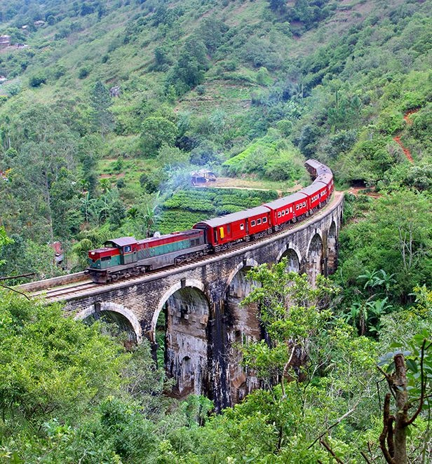 Red and green train crossing a stone arched railway bridge near Kandy, Sri Lanka, surrounded by lush green hillsides and dense forest.