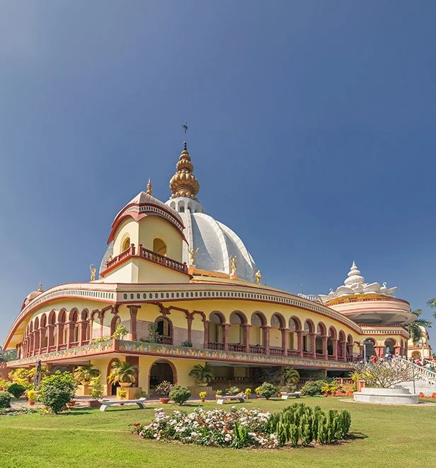 Ornate temple complex in Mayapur, India, with cream and red arched balconies, lush green gardens in the foreground, and a large white dome topped by a golden spire against a clear blue sky.