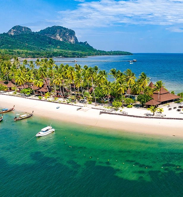Aerial view of Muk Island in Trang, Thailand, with a curve of white-sand beach lined with palm trees and turquoise water against a backdrop of green hills.