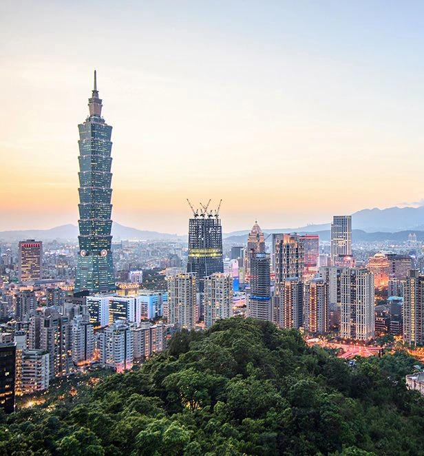 Evening view of Taipei’s skyline in Taiwan, with Taipei 101 rising above the city lights and a green forested hill in the foreground.