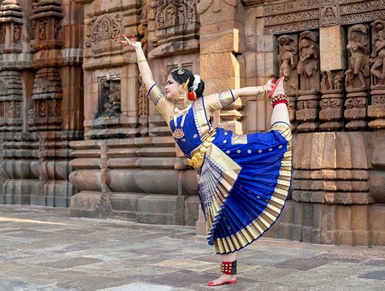 Bharatanatyam dancer in a vibrant blue and gold costume balancing in an elegant pose outside a carved stone temple, with one leg raised behind and arms extended gracefully.