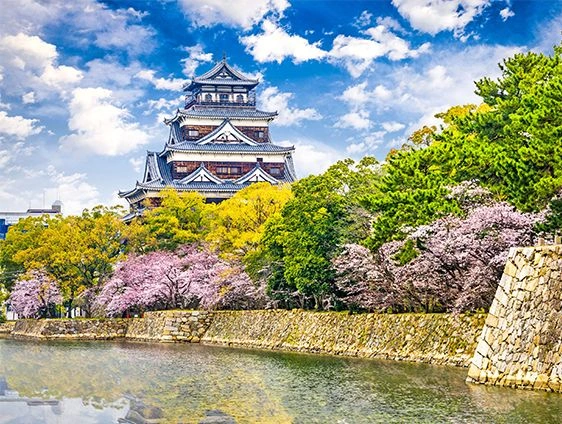 Hiroshima Castle rising above stone walls and a moat, framed by green trees and pink cherry blossoms in Japan.