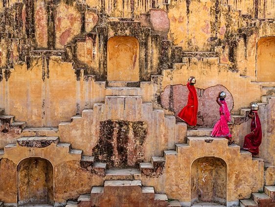 Three women in vibrant red and pink saris walking along the geometric steps and niches of an old sandstone stepwell in Jaipur, India, creating a striking pattern of colour against the weathered walls.