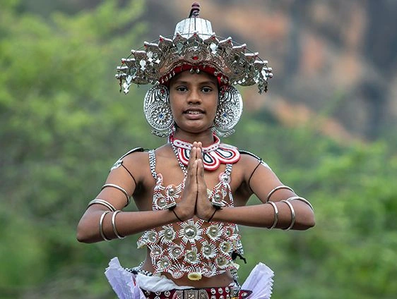 Kandyan dancer in Sri Lanka wearing elaborate silver headdress and traditional beaded costume, standing outdoors with hands pressed together in a greeting gesture against a blurred green backdrop.