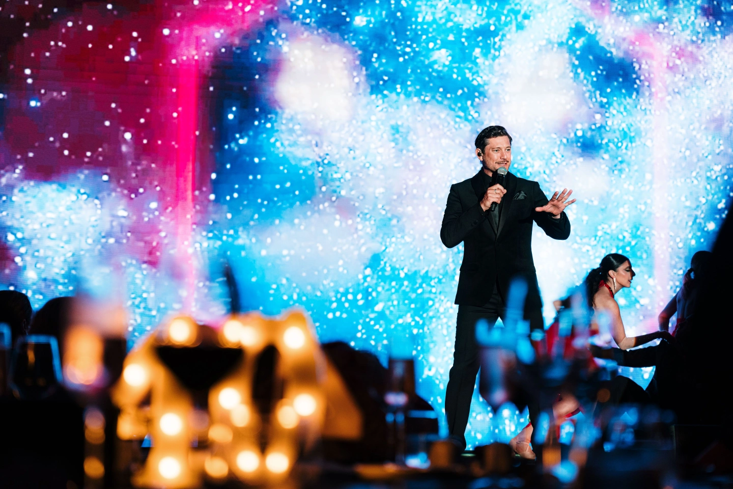 Male singer wearing a suit holding a microphone on a stage in front of a blue and pink screen