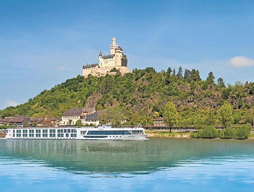 The Scenic Opal cruise ship on the Rhine River, with the backdrop of towering mountains and the historic Marksburg castle perched on top.