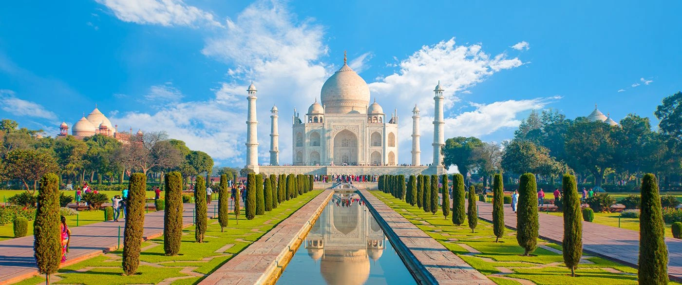 Taj Mahal in Agra reflected in the long central waterway, framed by formal gardens and cypress-lined paths, with visitors walking under a bright blue sky with scattered clouds.
