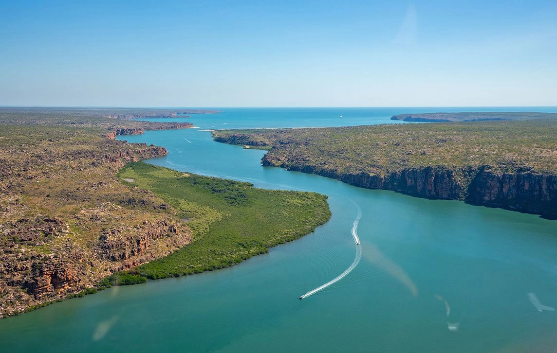 Aerial view of the King George River in the Kimberley, with a narrow boat tracing a white wake between rugged red cliffs and green mangrove-covered banks towards the open sea.