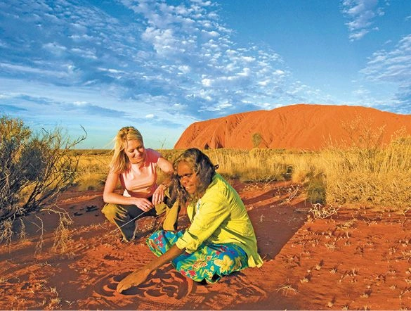 Caucasian woman crouching next to an Aboriginal woman in front of Uluru