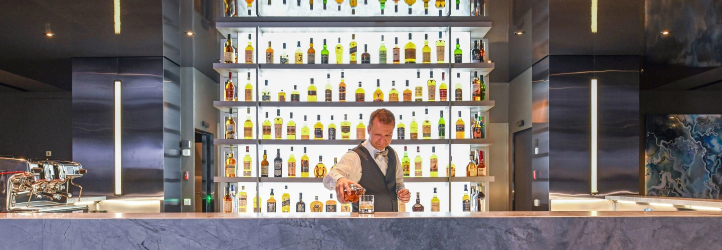 Bartender pouring whisky at a marble bar with a backlit wall of premium bottles