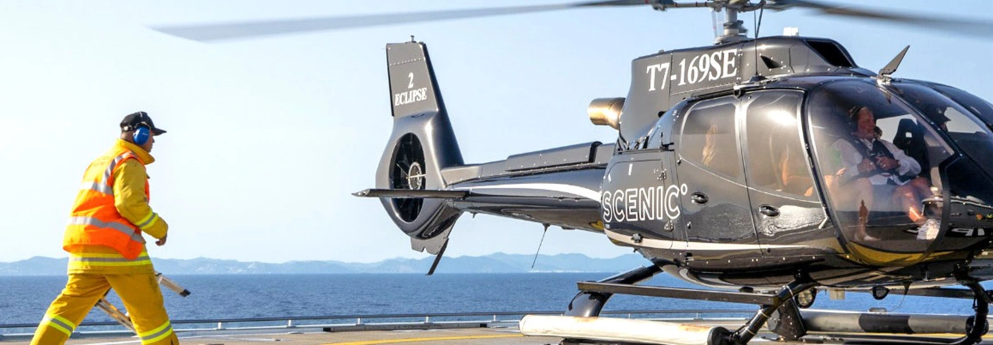 Scenic-branded helicopter preparing to take off from a ship’s helipad with safety crew nearby