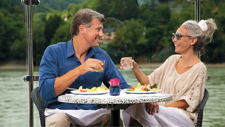 A man and woman smiling at each other with their drinks in hand as they eat lunch alfresco