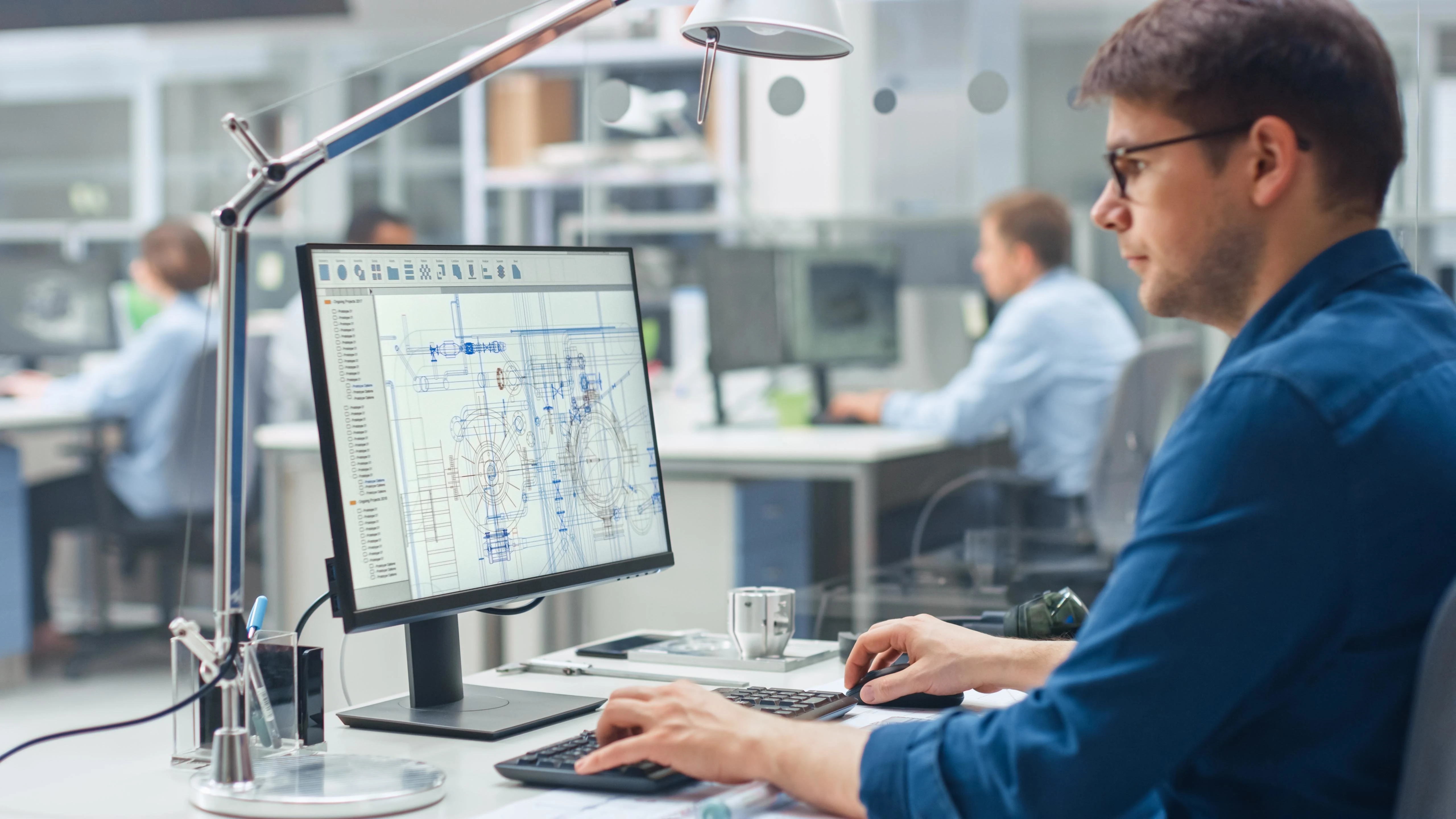 Engineer working at a desk in an open office, typing at a keyboard while viewing a technical CAD blueprint on a computer monitor