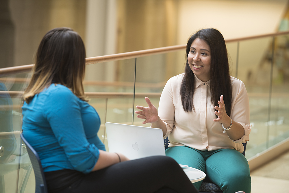 Two brown haired women sitting facing each other and talking with a laptop in between
