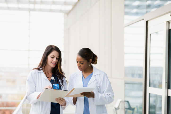 Two female nurse practitioners meet in a hospital hallway to discuss patient records.